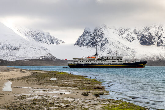 Passenger expedition ship sailing through Arctic fjord water beside a remote beach and massive snow-covered mountains, Spitsbergen, Svalbard, Norway