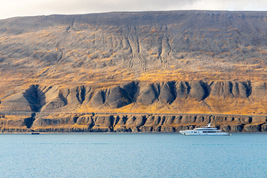 White yacht on calm Arctic fjord water beneath a vast barren mountain with deeply eroded slopes and layered terrain in summer, Spitsbergen, Svalbard, Norway