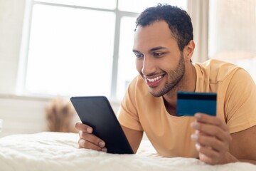Smiling Happy Young African American Man Shopping Online With Credit Card While Lying On Bed, Using Tablet At Home. Engaged In Online Shopping. Business Concept. © Prostock-studio