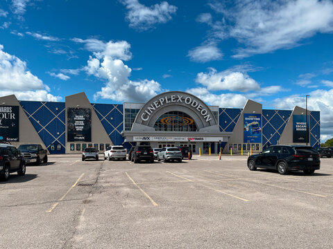 Wide shot of Cineplex Odeon cinema building with blue facade and arched entrance under blue sky with clouds and cars in parking lot, August 2026