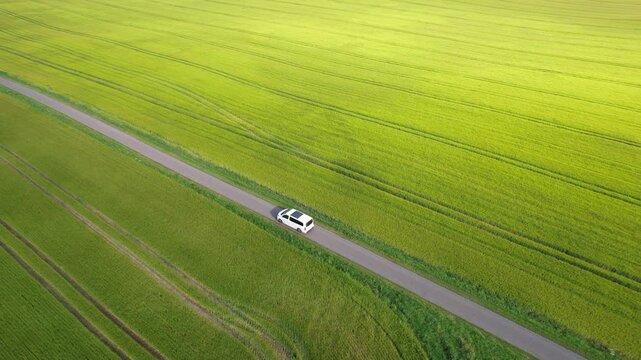 Aerial view of a white passenger minivan driving along a road through a green field.