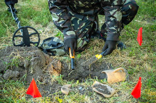 Camouflaged detectorist kneels beside soil plug, using handheld pinpointer to check target in grassy field. Metal detecting search feels focused and careful, with marked dig site and buried find