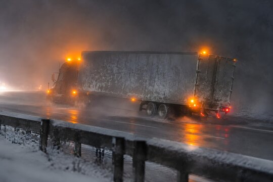 Jackknifed trailer angled across a highway lane on a winter night with salt spray and glowing emergency beacons