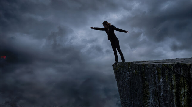 Businesswoman balancing on edge of high cliff under dark storm clouds. Corporate risk taking and overcoming challenges in hostile economic environment.