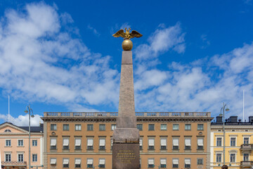 Fototapeta premium Empress Stone monument featuring a golden double-headed eagle statue atop an obelisk, standing prominently against a blue sky, surrounded by historic buildings in Market Square, Helsinki, Finland