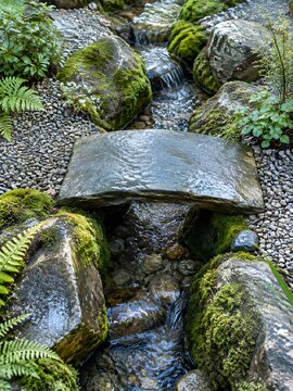 A fairy garden with a small flat stone bridge crossing a narrow trickle between mossy rocks on a gravel patio