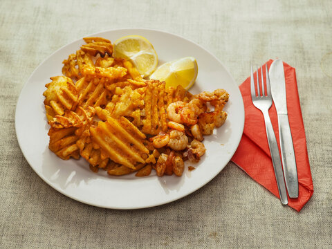 Plate holds crispy golden waffle fries and small shrimp. Two lemon wedges add freshness. Red napkin and silverware sit beside plate. Image shows appetizing seafood and fry combo