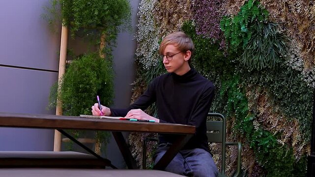 Young man wearing a black turtleneck and glasses passionately drawing or making notes with a pen in a sketchbook while sitting in a patio with green plants.