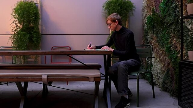Young man wearing a black turtleneck and glasses passionately drawing or making notes with a pen in a sketchbook while sitting in a patio with green plants.