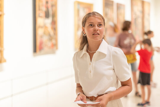 Young woman with sheet piece of paper in hands walks around museum hall, examines showpiece painting and icons at exhibition. List of recommended exhibitions, guidebook, and map of museum.