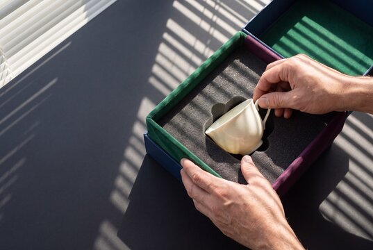 Hands carefully unpacking an antique porcelain teacup from a velvet lined luxury gift box on a sunlit table