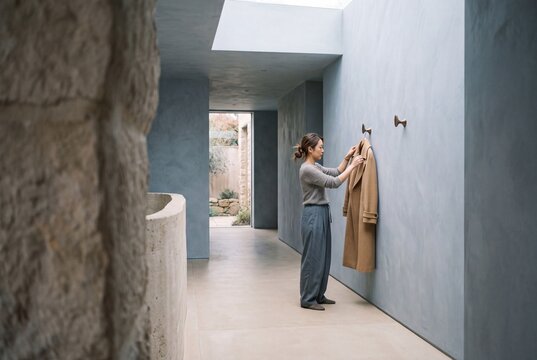 Asian woman hanging an elegant beige coat on a wooden wall hook in a modern minimalist hallway
