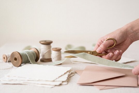 Woman hand cutting sage green velvet ribbon with vintage brass scissors for wedding stationery invitation design