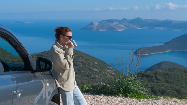 Relaxed woman drinking coffee on road trip enjoying scenic sea view. Happy adult woman leaning against her car during a travel stop, drinking from a mug and contemplating the coastal landscape