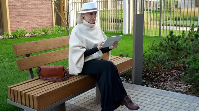 Woman with white hat reads tablet while resting on wooden bench. Tablet reading fills this peaceful garden moment, and her thoughtful expression stays fixed on handheld screen as handbag rests nearby.