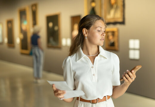 Museum female visitor with phone in hands walk through exhibition halls museum. Girl with guidebook inspects paintings of various artistic genres. Exposition, admires works of Renaissance artists