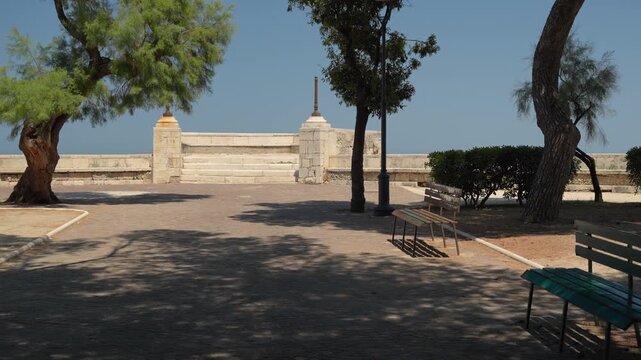 Green wooden bench standing under tamarisk trees in a sunlit seaside park in Trani, stone balustrade, lamp posts and clear blue sky behind, concept of relaxation, public space and slow summer days