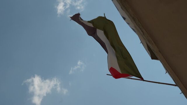 Low angle view of a Palestinian flag waving on a pole mounted to building wall against blue sky with scattered white clouds, concept of national symbols, identity and international politics