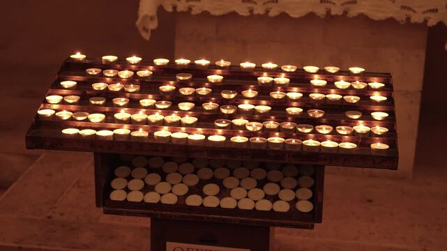 Rows of small votive candles burning on a dark metal stand inside a church, unlit tea lights waiting on the lower tray, warm flickering glow in dim light, concept of prayer, remembrance and faith