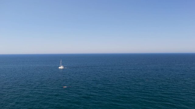 Small white boat floating alone on calm deep blue sea beneath a wide pale blue sky, a red buoy bobbing nearby and the straight horizon splitting the view in half, concept of simple life and quiet esc