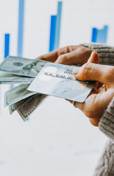 Hands holding US dollars currency and a bank card in front of the stock exchange screen showing trading graphics.