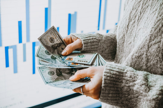 Hands holding US dollars currency in front of the stock exchange screen showing trading graphics.