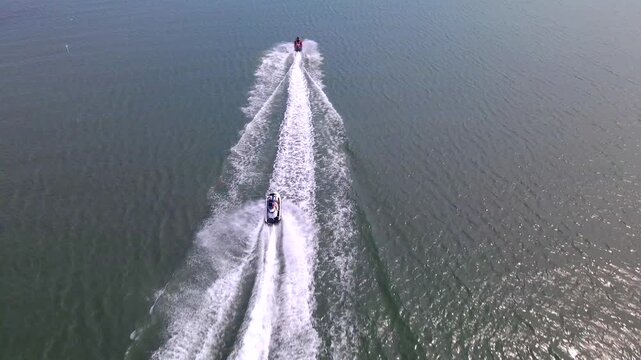 Overhead drone view follows two jet skis cutting foamy trails through green water.
