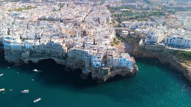 In bright sunlight, the white clifftop town of Polignano a Mare rises from a rocky promontory above deep blue sea, sparkling water and scattered boats in the foreground, concept of summer escape and 