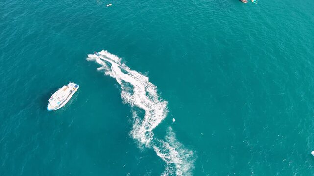 Speeding boat carving a winding white wake across deep teal sea, seen from directly above. Motor yachts cruise nearby on open water, concept of luxury boating, speed and summer leisure on the coast