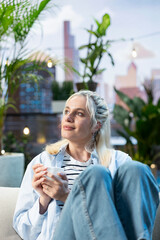 Obraz premium Relaxed woman holds a white coffee cup and looks upward while daydreaming on a rooftop sofa, enjoying a peaceful break in a modern urban outdoor lounge.