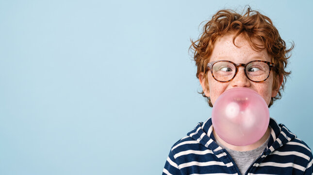 Red haired freckled boy with glasses crossing his eyes as he blows a huge pink bubble gum bubble, playful, cheeky childhood portrait on blue background with copy space