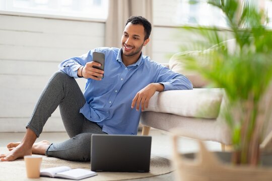 Happy Male, Young Adult, Black, Sitting On Floor With Laptop And Smartphone At Home, Engaged In Work Or Communication. Comfortable Setting Indicating Remote Work Lifestyle, Business Concept.
