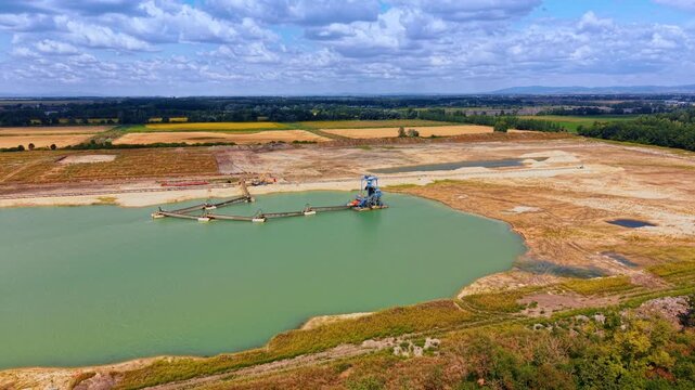 Sand dredging equipment operating in a quarry pond. Industrial machinery works in green water surrounded by flat agricultural fields under clouds.