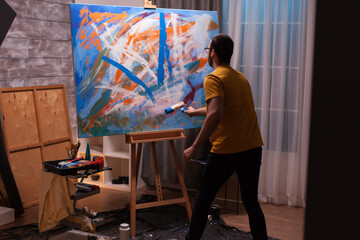Male artist uses a paint roller to add sweeping blue marks to a large abstract canvas, combining energetic movement, experimentation and expressive creativity. © DC Studio