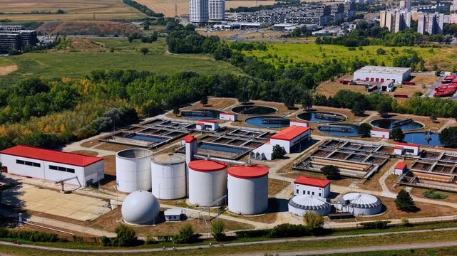 Cylindrical tanks at a wastewater treatment plant. Four white cylinders stand beside a domed tank, with settling basins and apartment buildings behind.