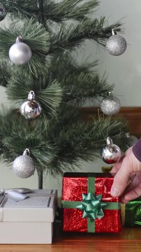 Man's hand taking a wrapped gift from under a Christmas tree decorated with silver balls, static shot with front view