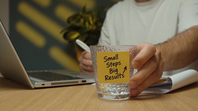 Man takes a sip of water, puts the glass with a &ldquo;Small Steps Big Results&rdquo; motivational sticky note back on the desk and continues working on his laptop.