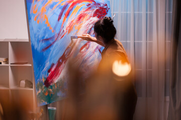 Woman artist leans toward a large canvas and works into the layered abstract surface with a brush, combining close attention to detail with focused brushwork and the creative process. © DC Studio