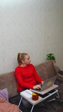 Young woman sits comfortably on sofa using laptop on portable lap desk at home. Relaxed home workspace includes notebook mug, tablet and soft furnishings in cozy interior. Suitable for remote work
