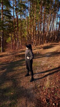 Young adult woman performs side lunge stretch during outdoor fitness session. Person wears black hoodie, cap, sweatpants and boots while warming up on grassy ground surrounded by autumn trees in