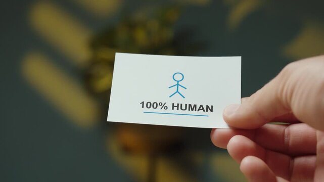 Close-up of a hand holding a &ldquo;100% Human&rdquo; card with a person symbol, representing human verification, authenticity and distinction between humans and artificial intelligence.