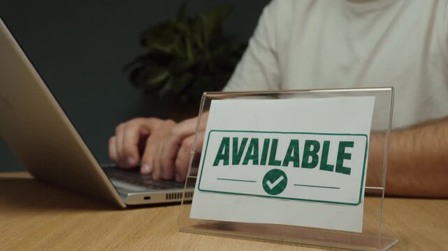 &ldquo;Available&rdquo; sign displayed prominently on a desk while a man works on a laptop in the background, representing availability, open service and readiness for business.