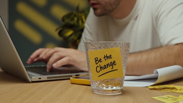 Man takes a sip of water, puts the glass with a &ldquo;Be the Change&rdquo; motivational sticky note back on the desk and continues working on his laptop.