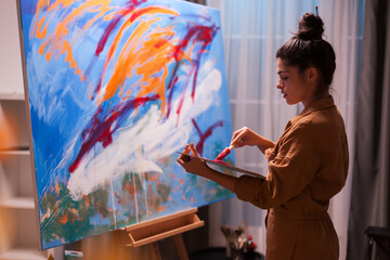 Woman artist dips a brush into red paint on a handheld palette beside an abstract canvas, a practical preparation step in an active studio painting session. © DC Studio