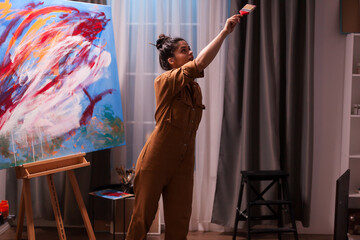 Painter looks upward while reaching high with a paintbrush beside a colorful abstract canvas, capturing movement and creative experimentation in the studio, highlighting artistic expression. © DC Studio