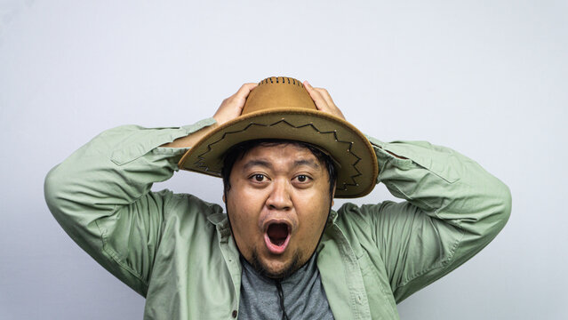 Showcasing extreme surprise, this young Asian male grips his brown cowboy hat using both hands while standing before some plain light background. The individual displays an incredibly