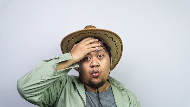 Experiencing sudden relief, one young Asian male wipes his forehead using one hand while sporting a brown cowboy hat against plain light background. The person features an expressive