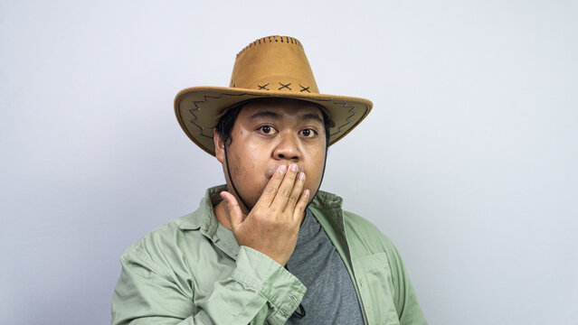Showcasing intense shock, a young Asian male covers his mouth using one hand while wearing western headwear plus green casual clothing before plain light background. The subject features