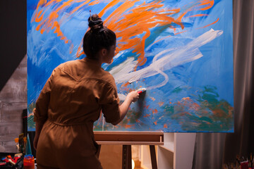 Woman artist paints flowing white curves over a blue and orange abstract canvas, shaping a layered contemporary composition with broad brushwork. © DC Studio