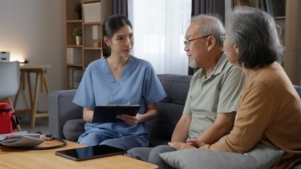 Obraz premium Professional Asian female nurse listening attentively to senior couple sitting on couch, holding medical chart during home healthcare visit in living room.